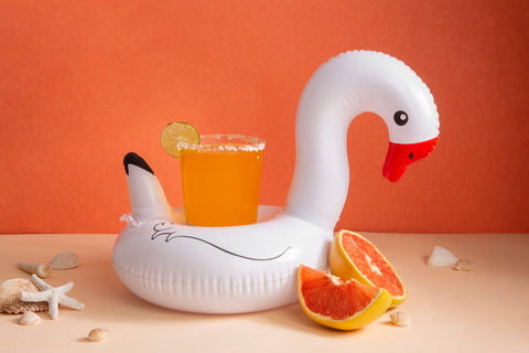 Refreshing summer drink in an inflatable swan cup holder surrounded by pool supplies like seashells and citrus fruit, perfect for a backyard pool day.
